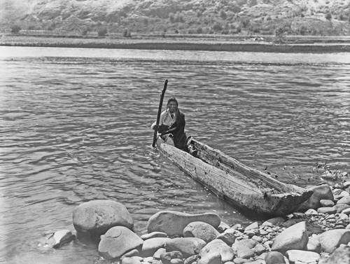 A Nez Perce man poling a small dugout canoe to shore